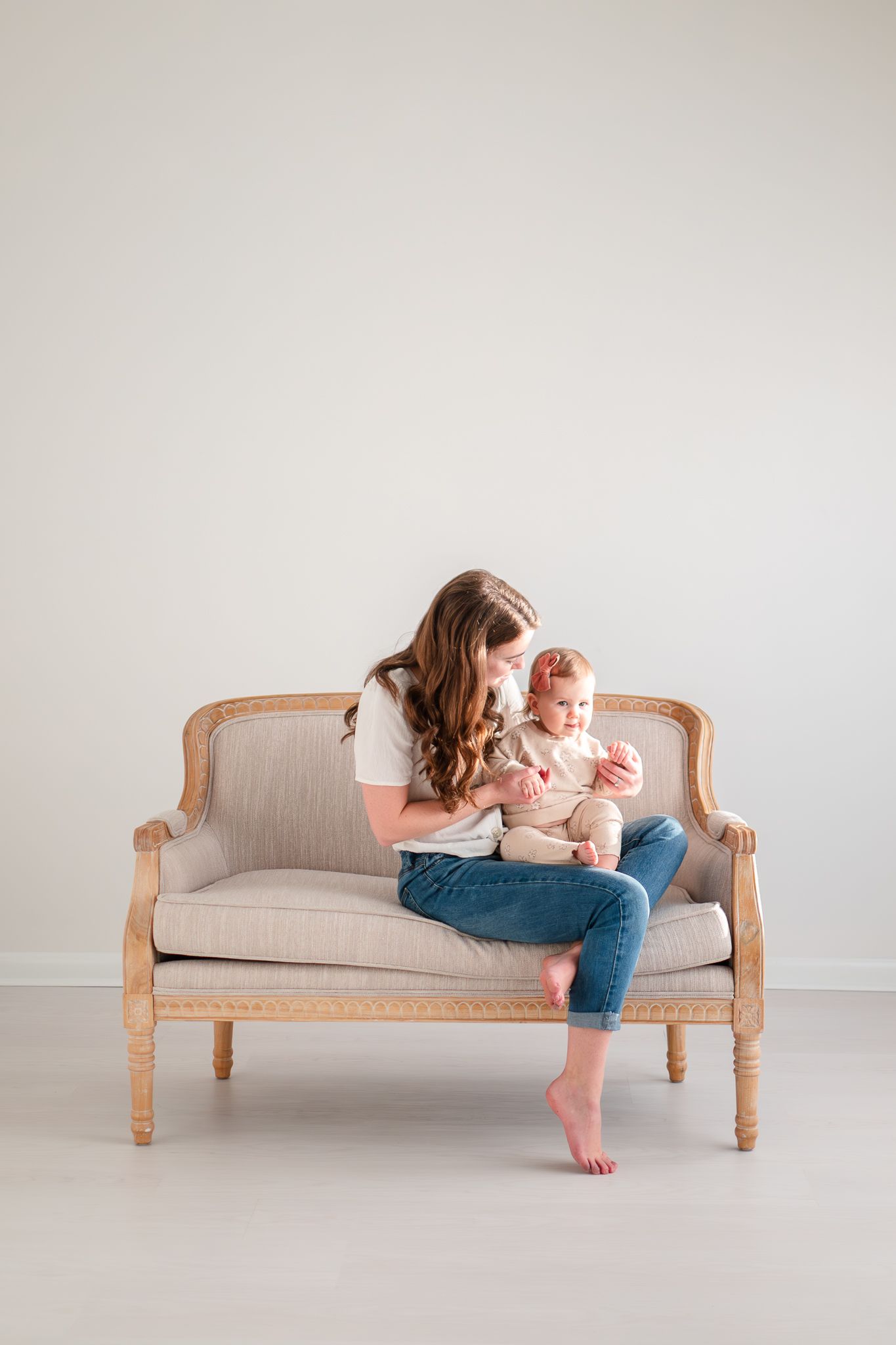 Mother and baby sharing tender moment while sitting on elegant beige loveseat with wood frame against neutral wall.