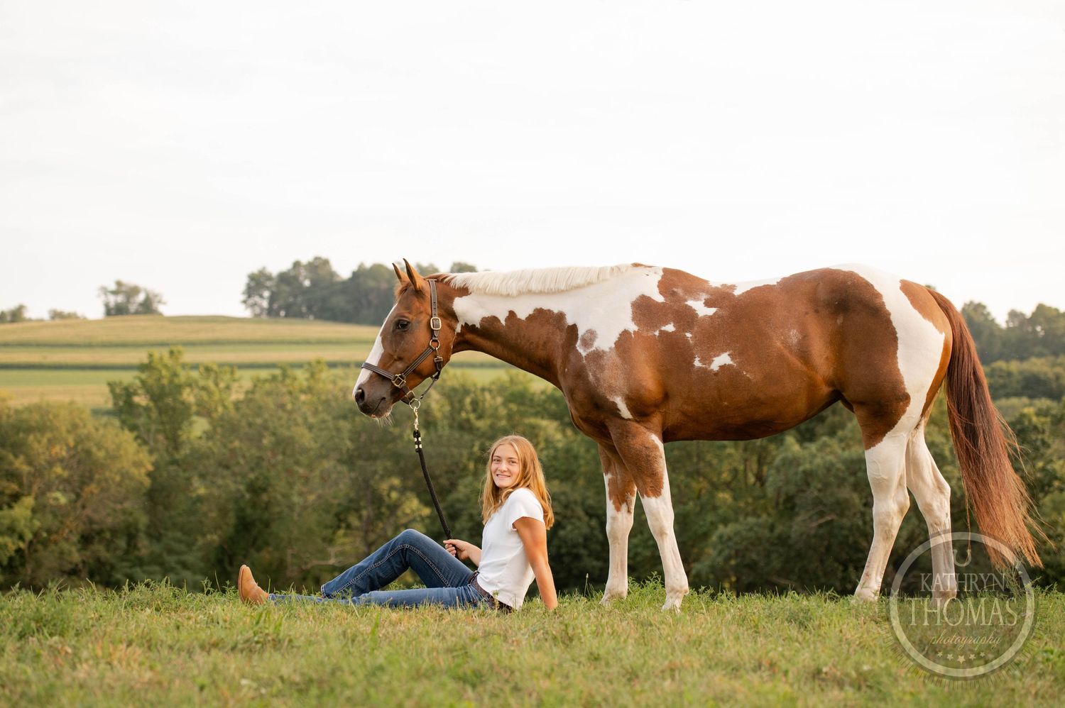Person relaxes in grass beside paint horse in serene country setting.