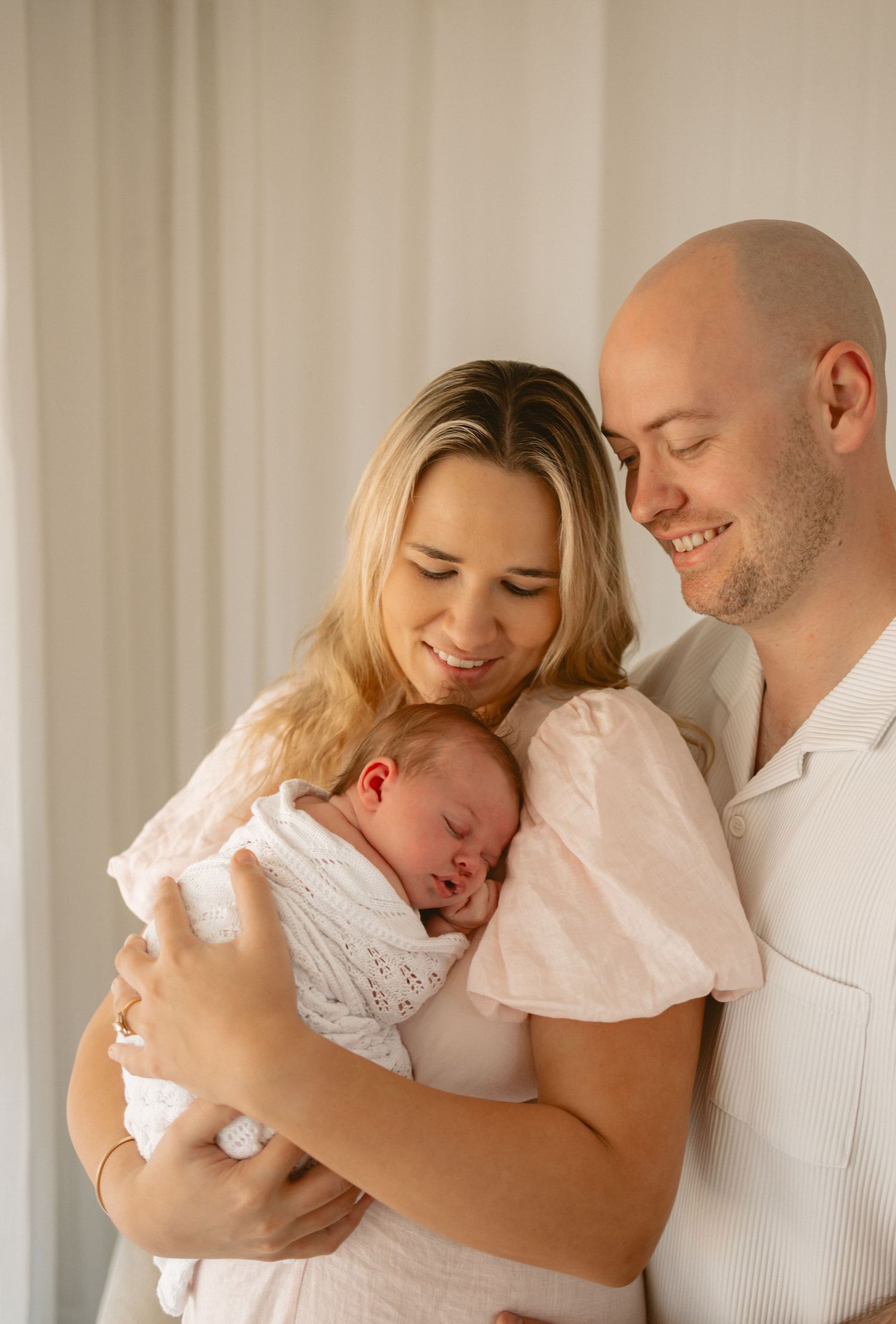 A family lovingly embraces their sleeping baby in a light-filled room wearing white clothing.