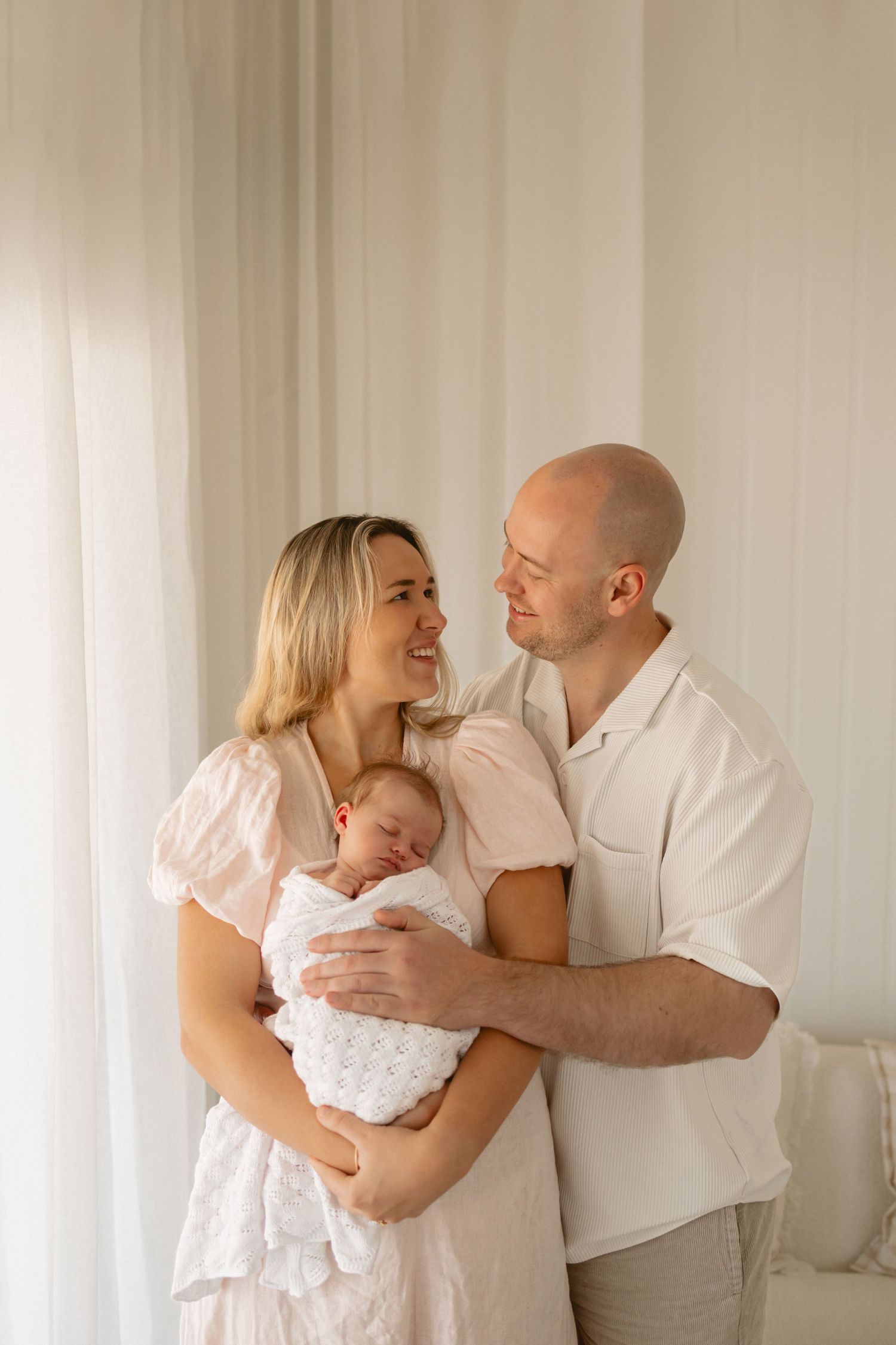 A warm family portrait series in soft white tones shows parents lovingly embracing their baby in a bright studio setting.