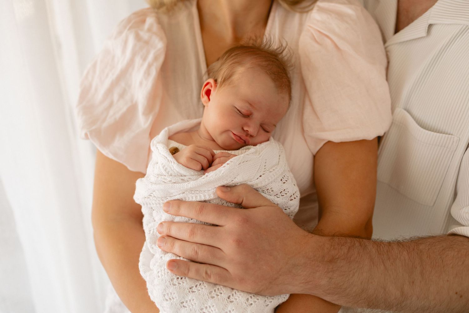 Adorable newborn baby sleeps peacefully in their parent's arms while wearing soft white clothing.