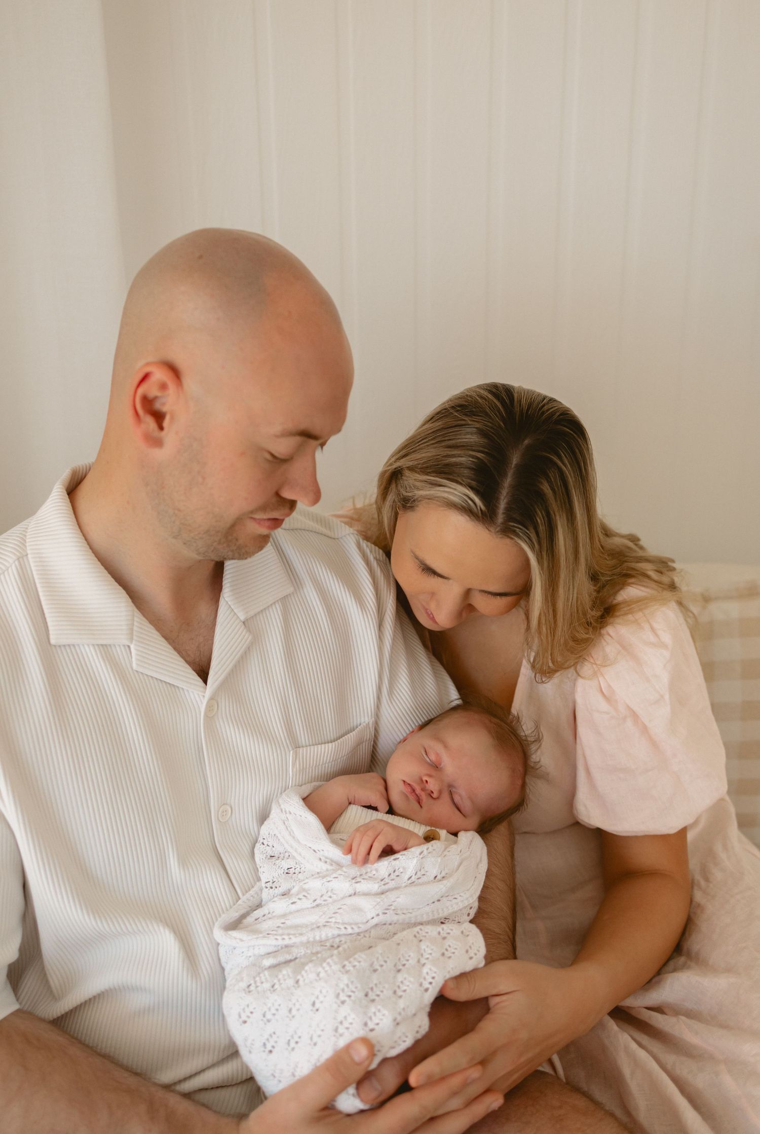 Parents in white and pink outfits tenderly gazing at their newborn baby wrapped in a knit blanket during a photo session.