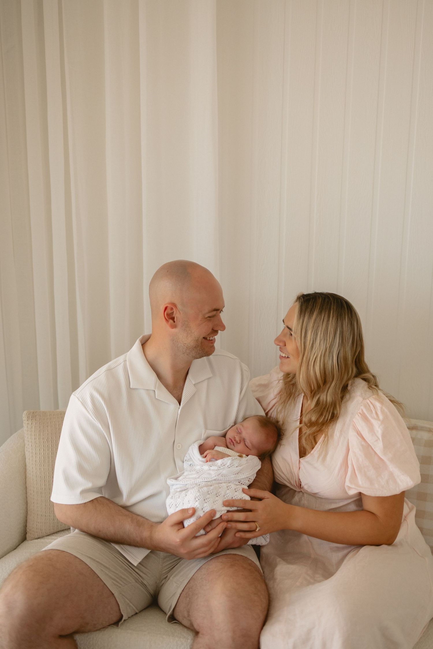 A couple in light colored clothing sits on a couch holding their newborn baby wrapped in white.