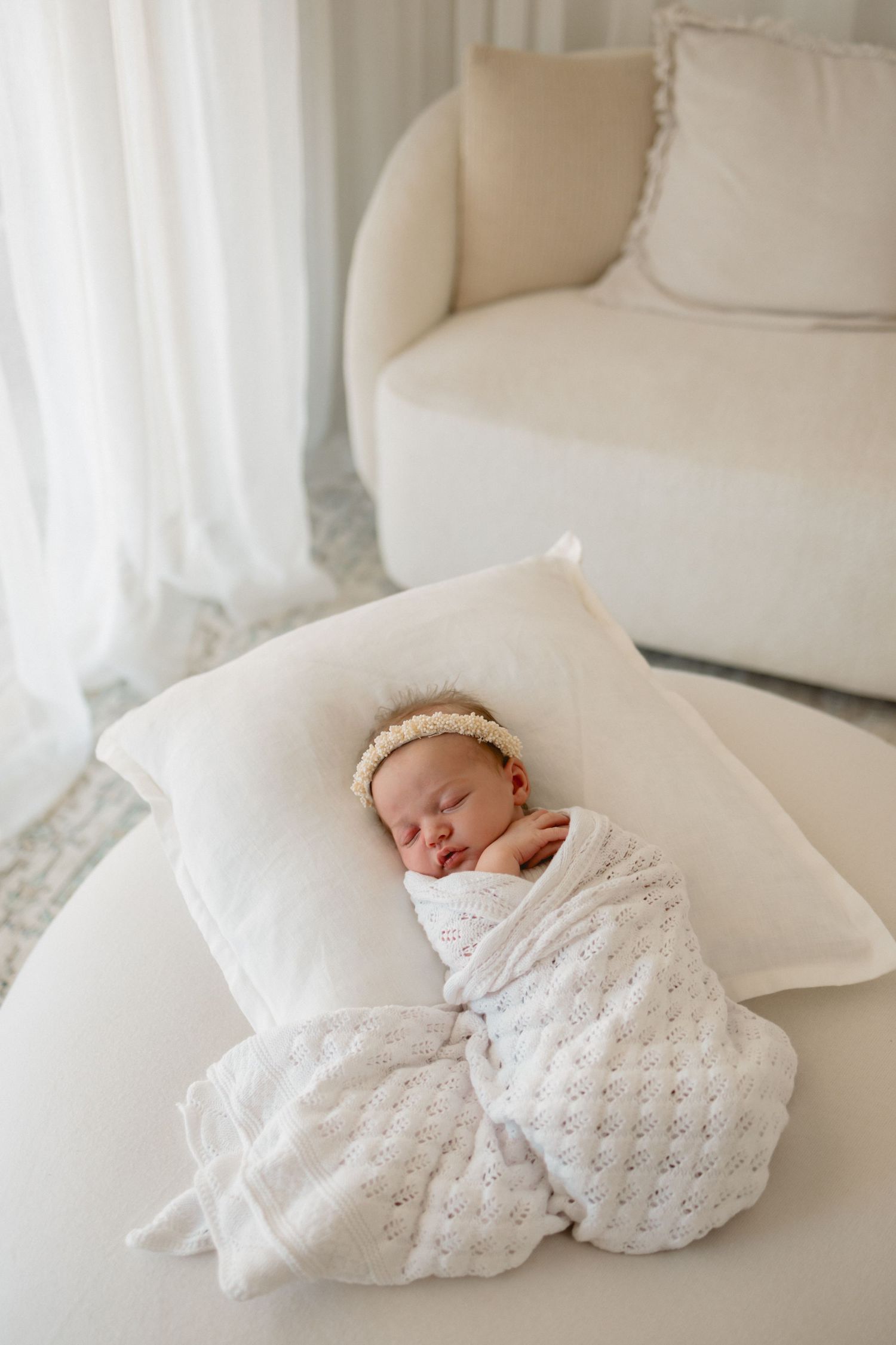 A newborn baby wrapped in delicate white lace swaddle rests peacefully on a white couch in natural light.