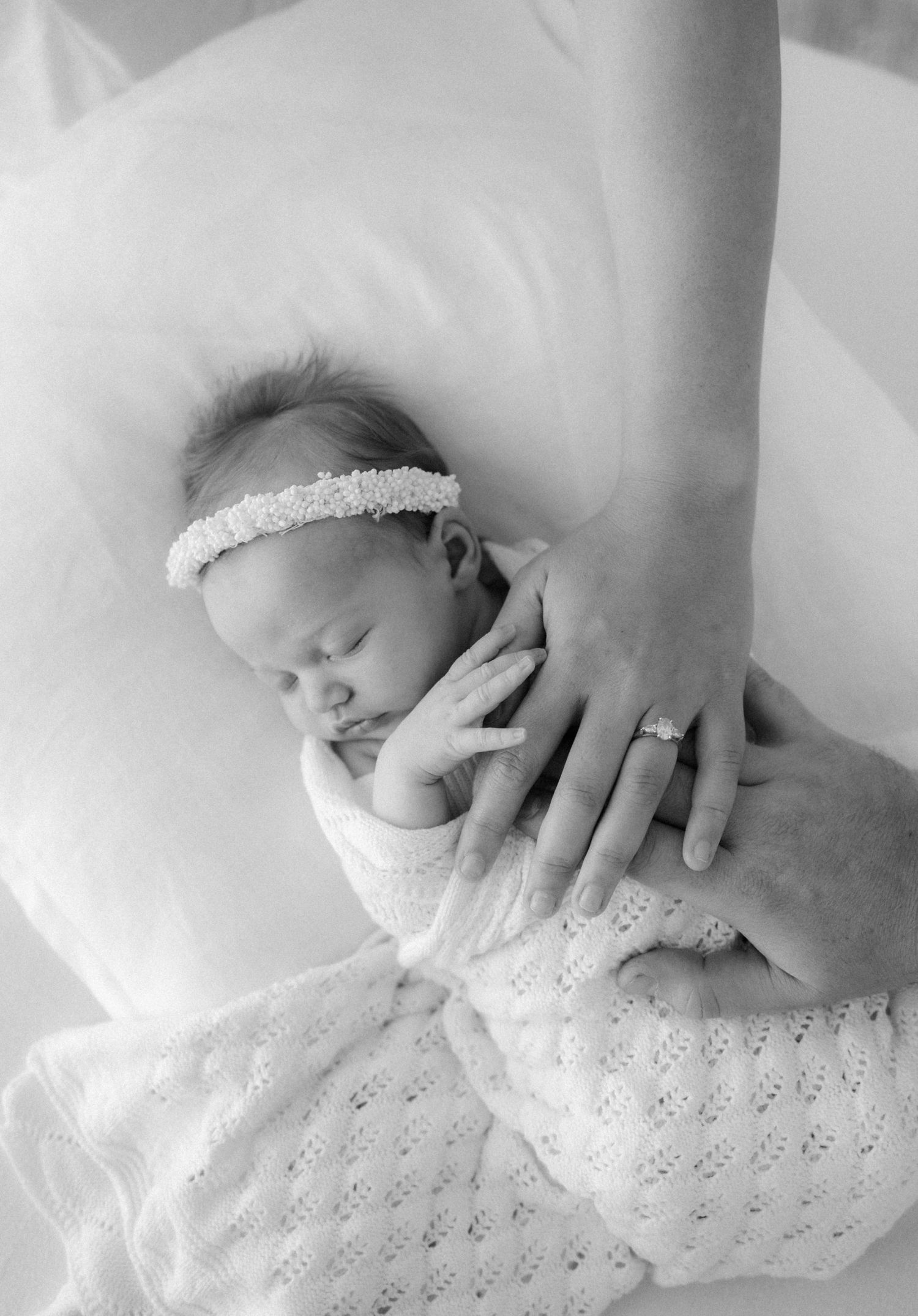 A newborn baby swaddled in white lace sleeps peacefully while someone's gentle hand rests on their head.