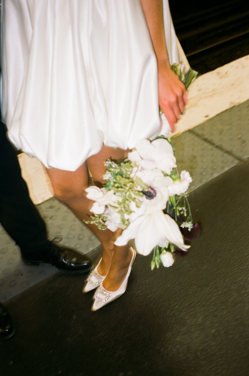 A bride in white shoes holds a delicate bouquet of white flowers against her dress while standing on pavement.