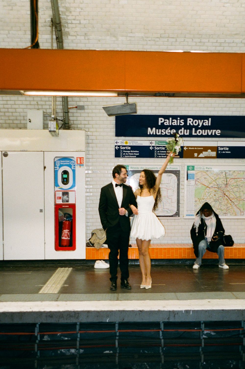 A couple in formal attire stands on a Paris metro platform at the Palais Royal-Musee du Louvre station.