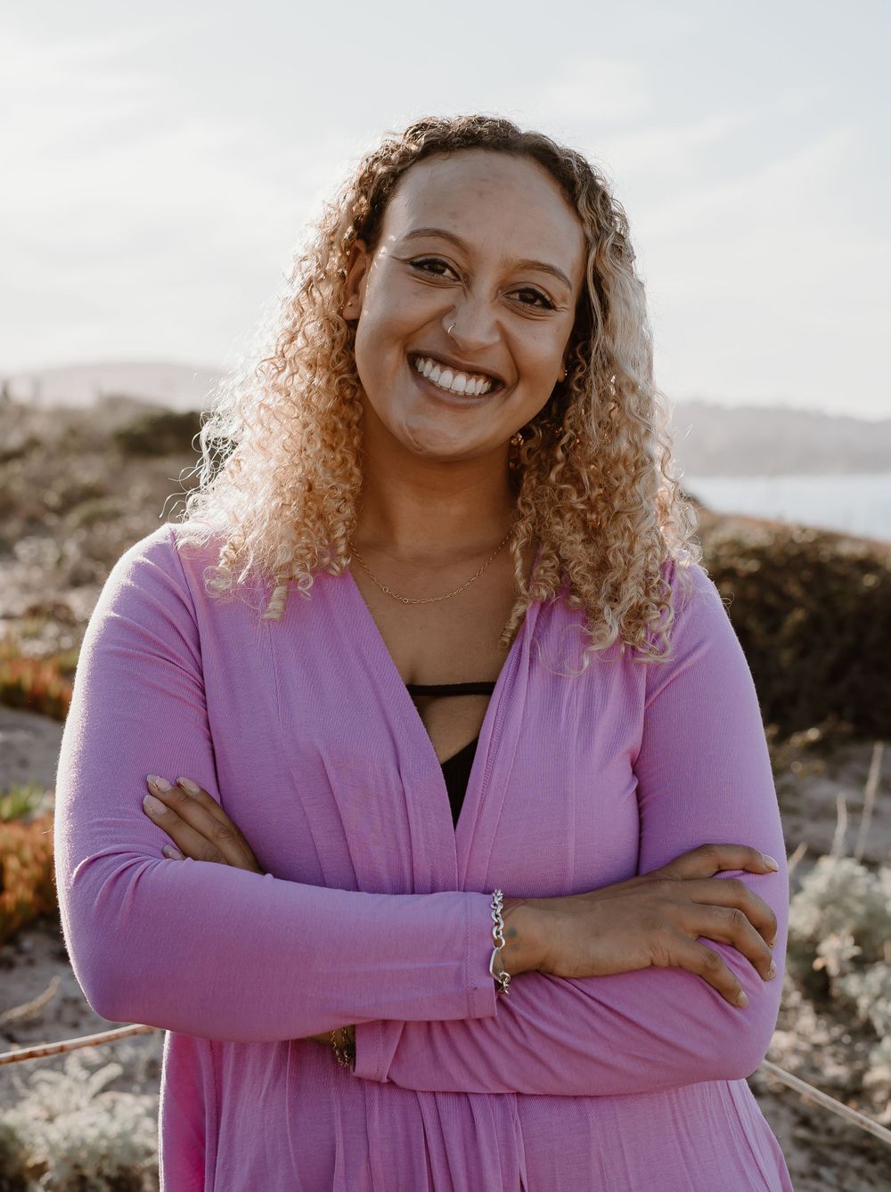 A person with curly hair and a purple cardigan smiles warmly while posing outdoors on a rocky coastline at sunset.