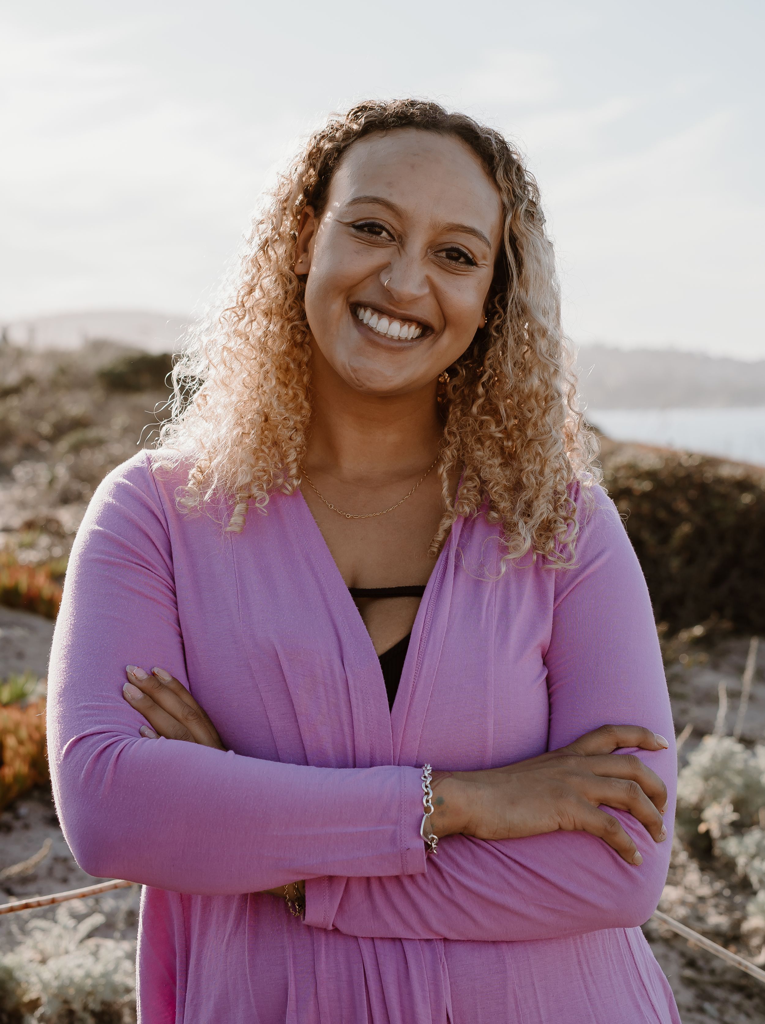 A person with curly hair and a purple cardigan smiles warmly while posing outdoors on a rocky coastline at sunset.