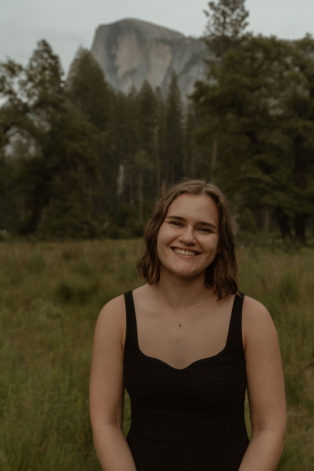 A person in a black tank top smiles warmly while standing in a meadow with Half Dome at Yosemite visible in the background.