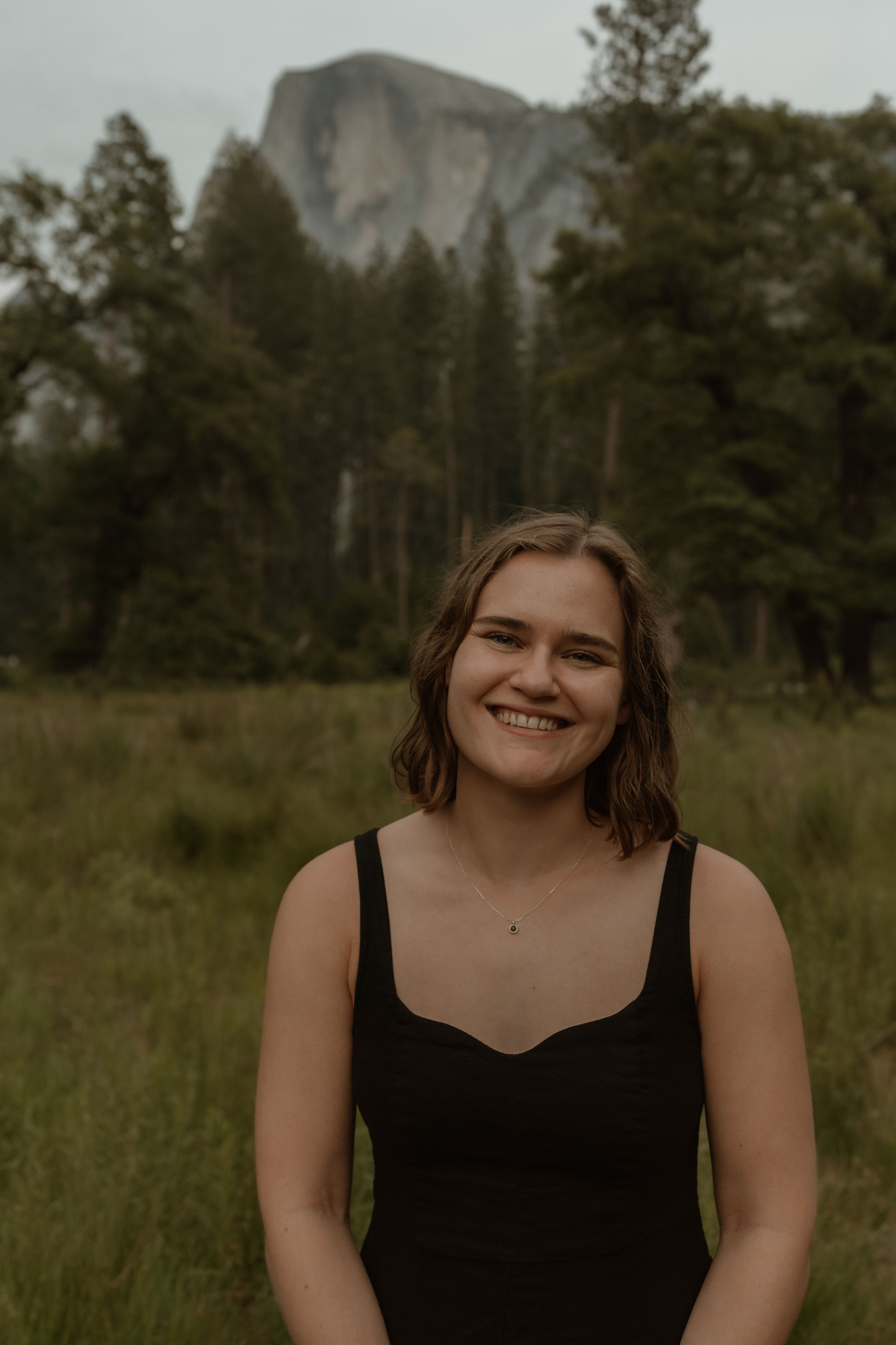 A person in a black tank top smiles warmly while standing in a meadow with Half Dome at Yosemite visible in the background.