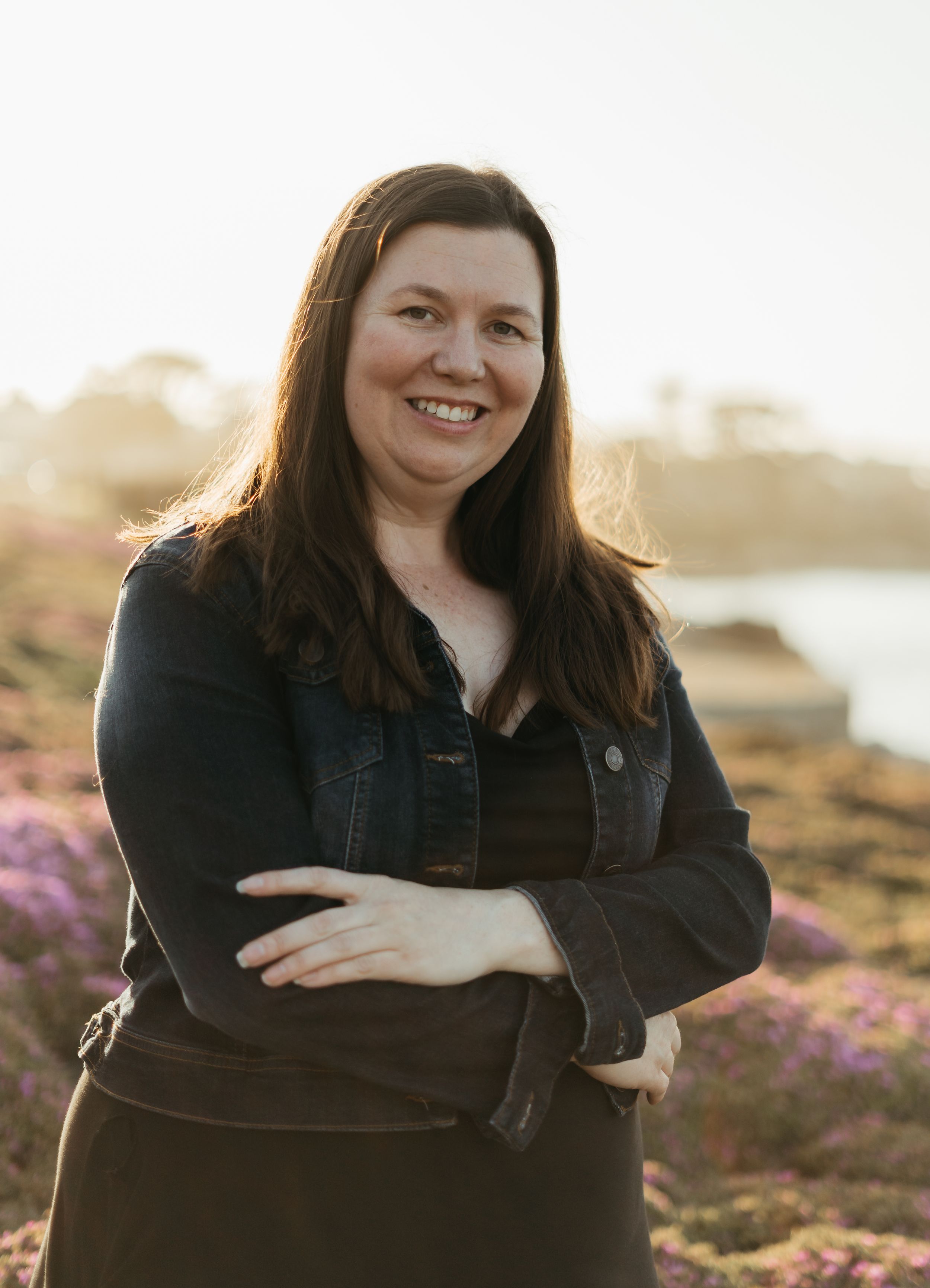 Person in dark attire poses outdoors against sunset backdrop in nature with purple flowers visible.