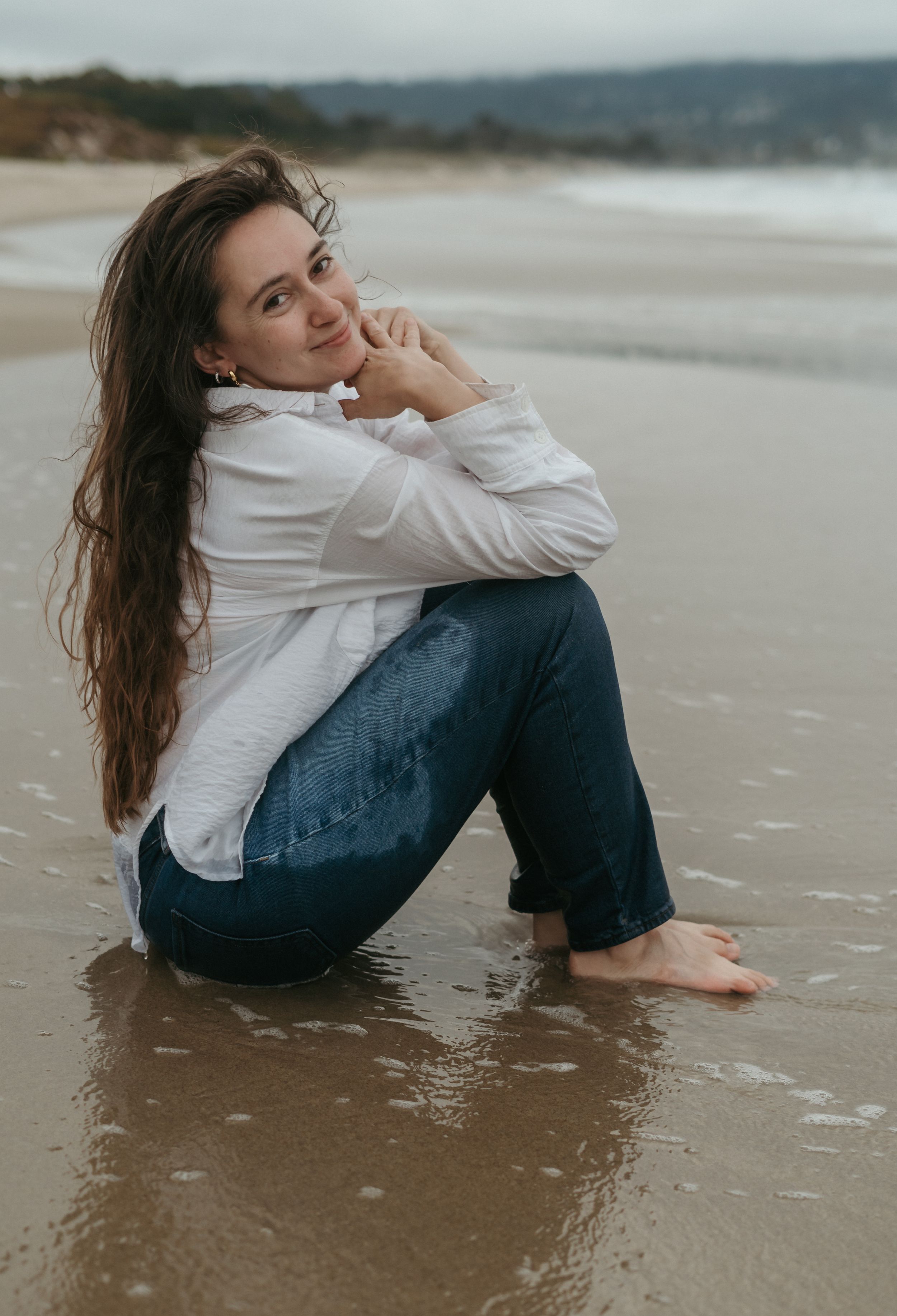 Someone in a white shirt and jeans sits reflectively on a sandy beach near gentle waves during overcast weather.