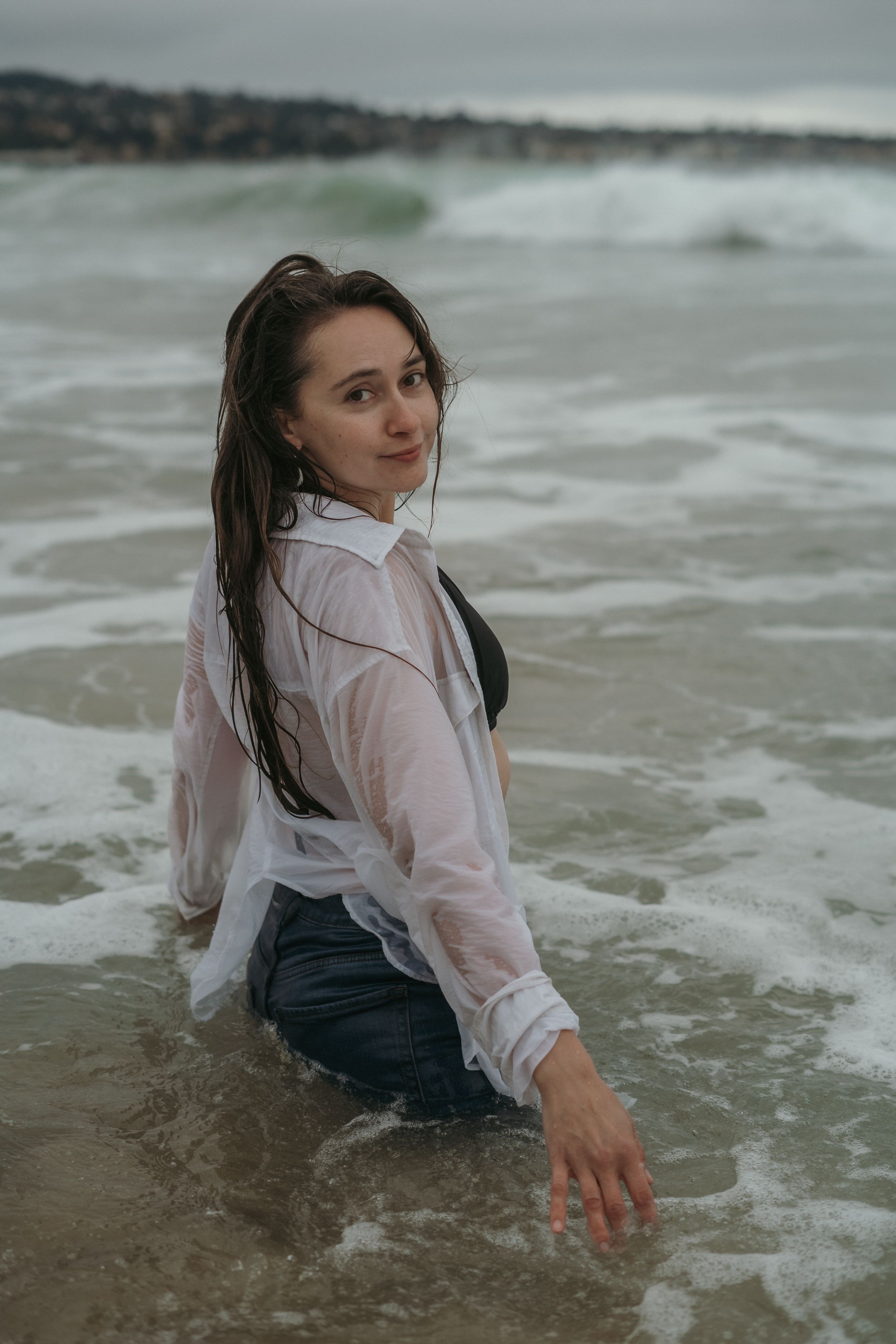 Someone wearing a wet white button-down shirt and dark pants sits in the shallow ocean waves at the beach.