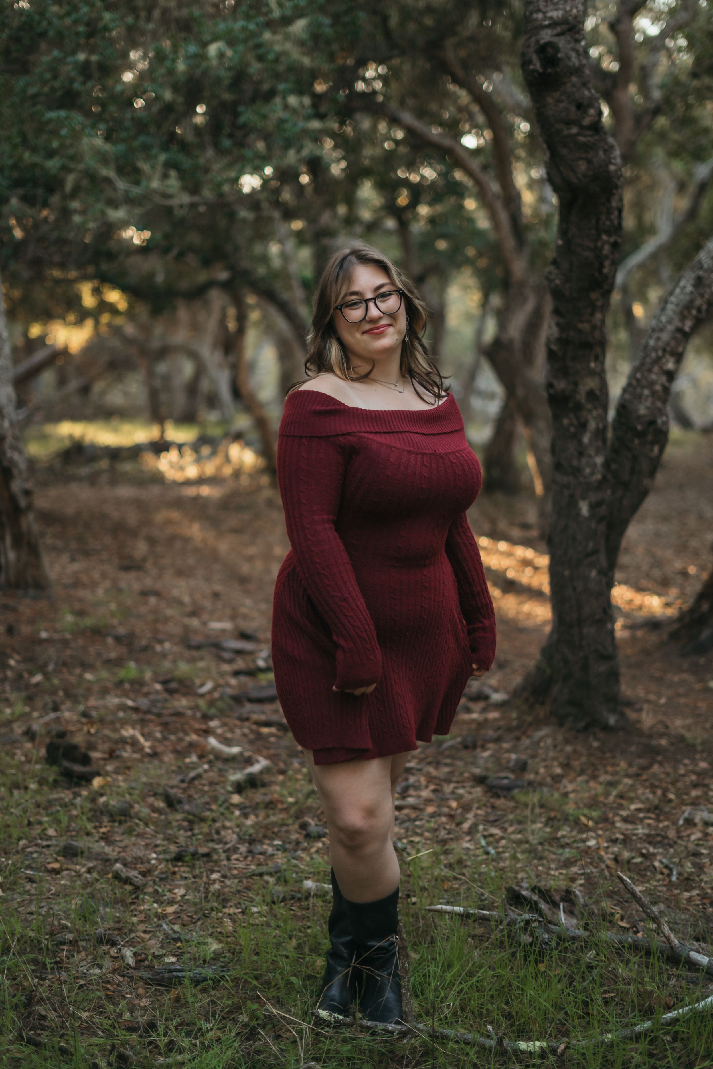 Person in a burgundy sweater dress poses among trees in a wooded outdoor setting during golden hour.