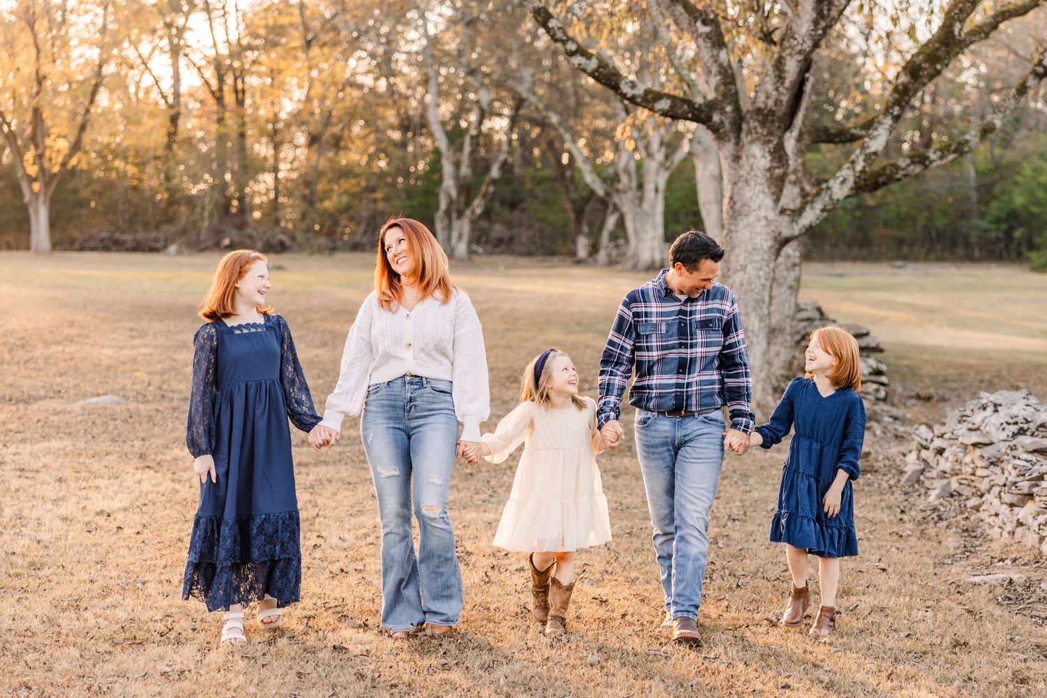 A family holding hands while walking together in a fall field at sunset wearing coordinating navy and denim outfits.