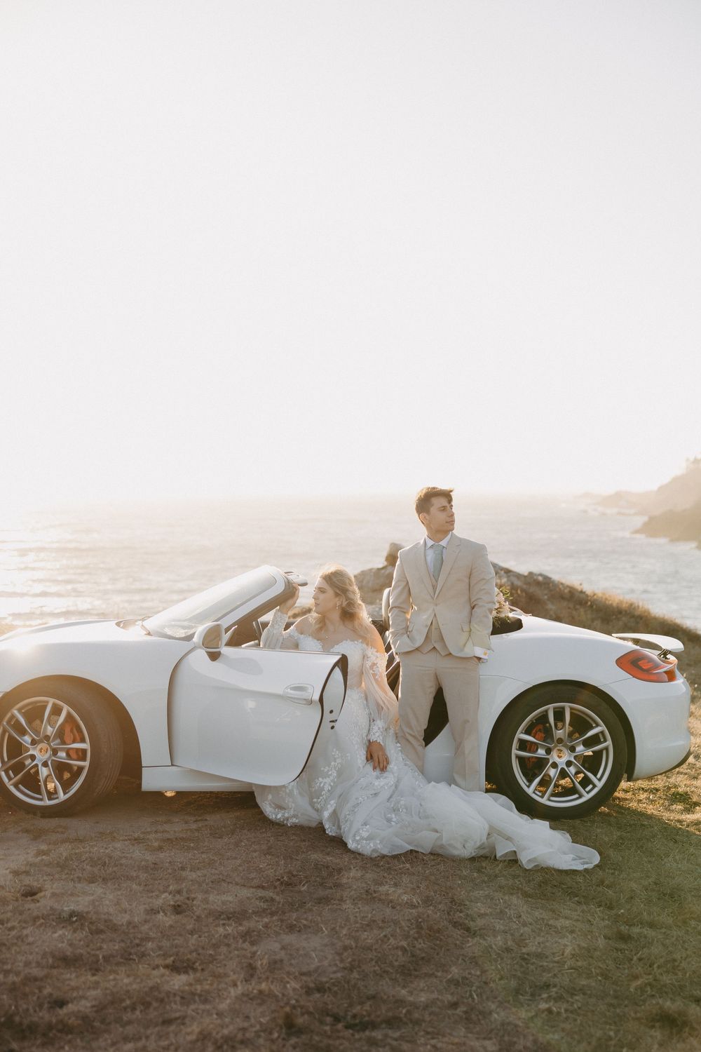 Newlyweds pose with a white luxury convertible sports car against a scenic coastal backdrop at sunset.