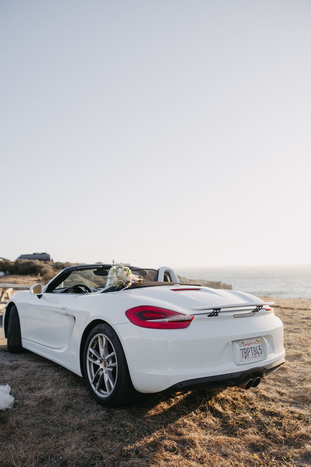 White Porsche Boxster convertible parked on a cliff overlooking the ocean during sunset.