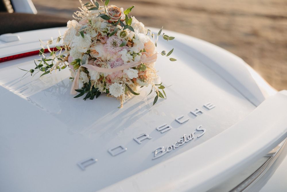 Small white and blush floral bouquet rests on a white Porsche car hood in natural lighting.