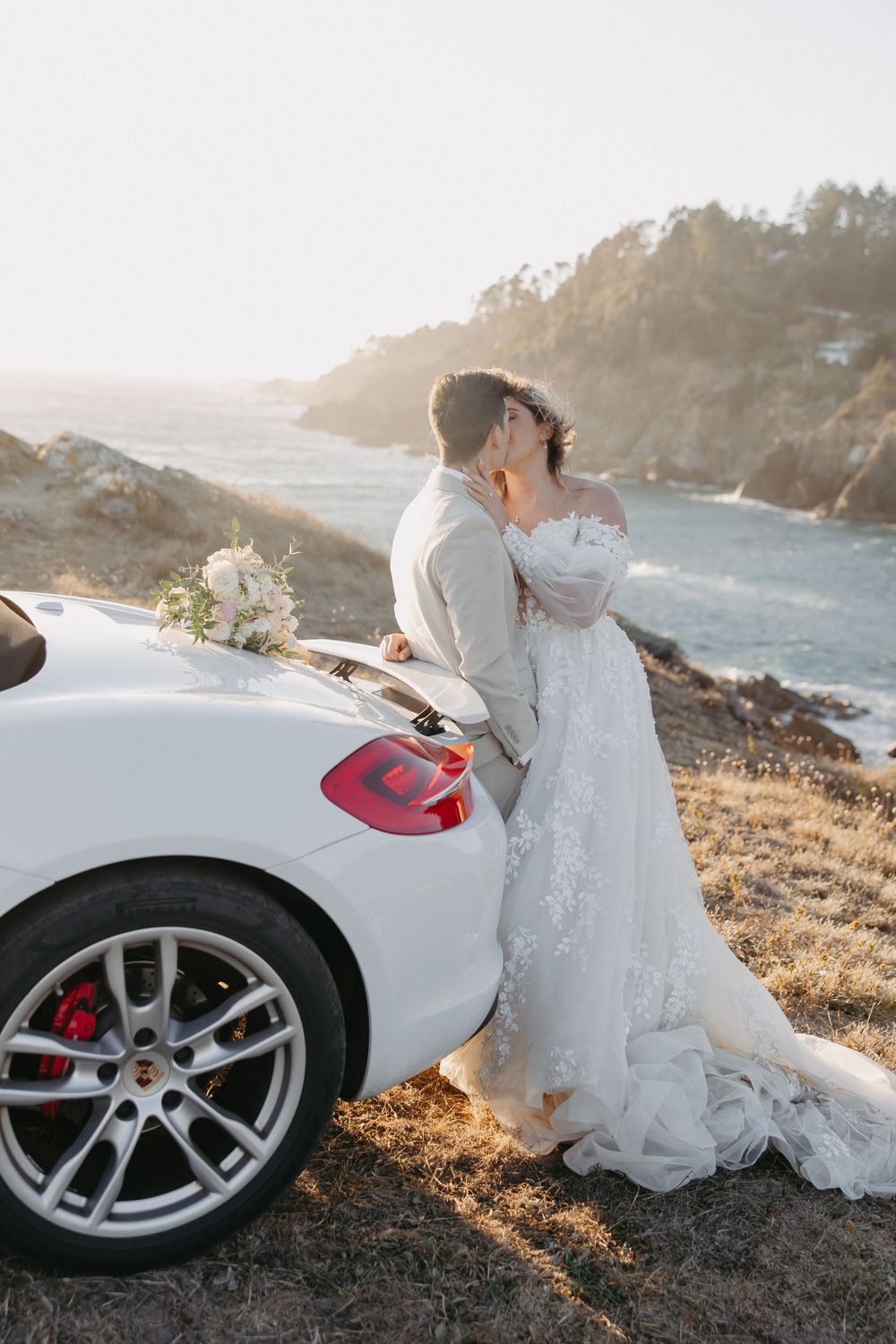 Wedding couple sharing an intimate moment beside a white sports car on a coastal cliff.