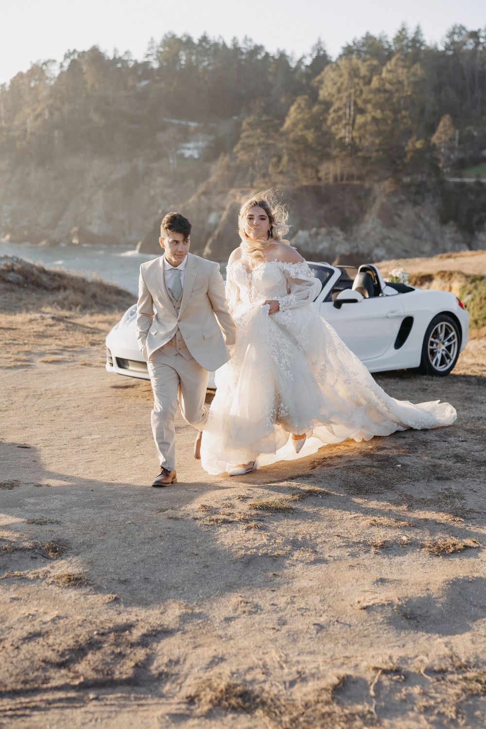 A couple in wedding attire running joyfully along a coastal path with a white sports car in background.