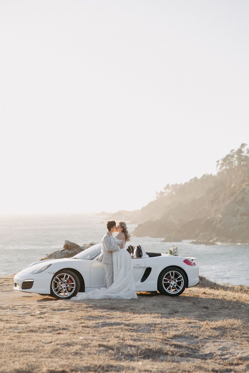 A elegant white wedding photo with a luxury Porsche sports car against a misty coastal cliff backdrop.