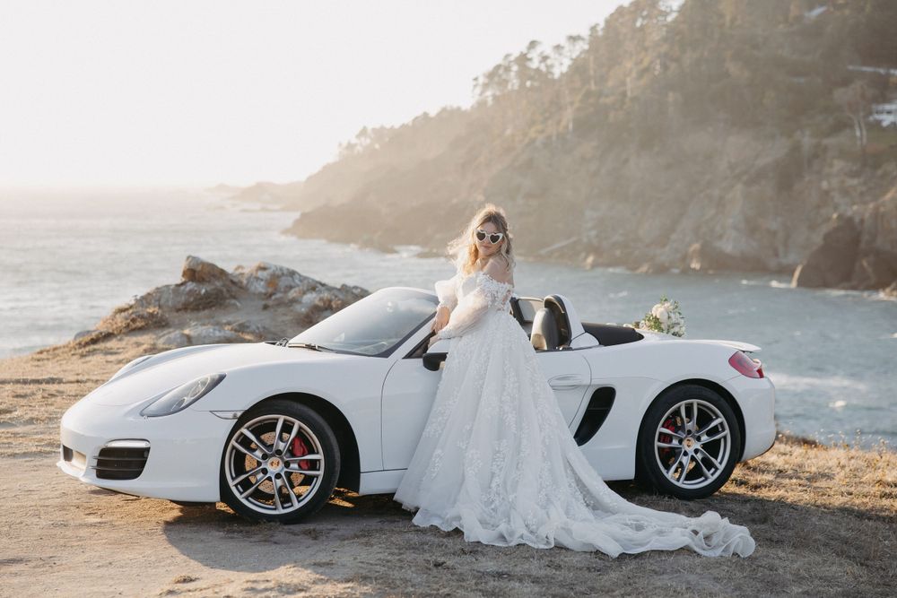 Luxurious white Porsche Boxster parked on scenic coastal cliff during sunset with figure in flowing wedding gown.