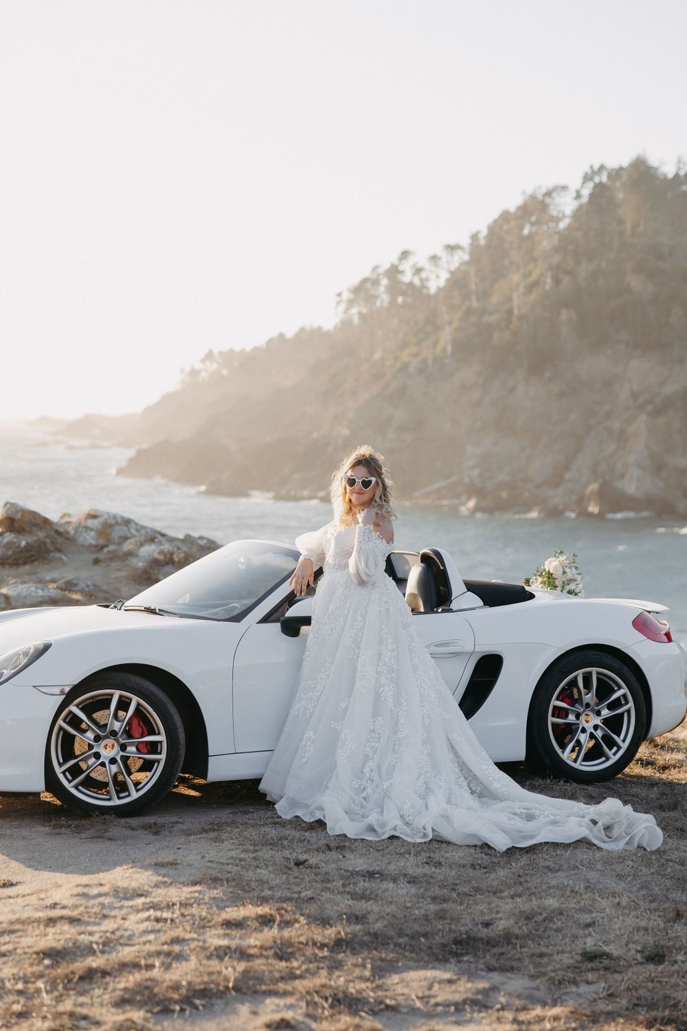A bride in a flowing lace gown poses beside a white Porsche convertible on a scenic coastal cliff at sunset.