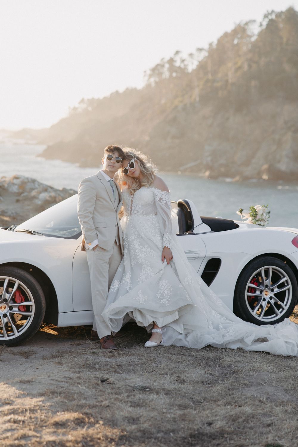 Newlyweds pose against their white Porsche convertible on a scenic coastal overlook.