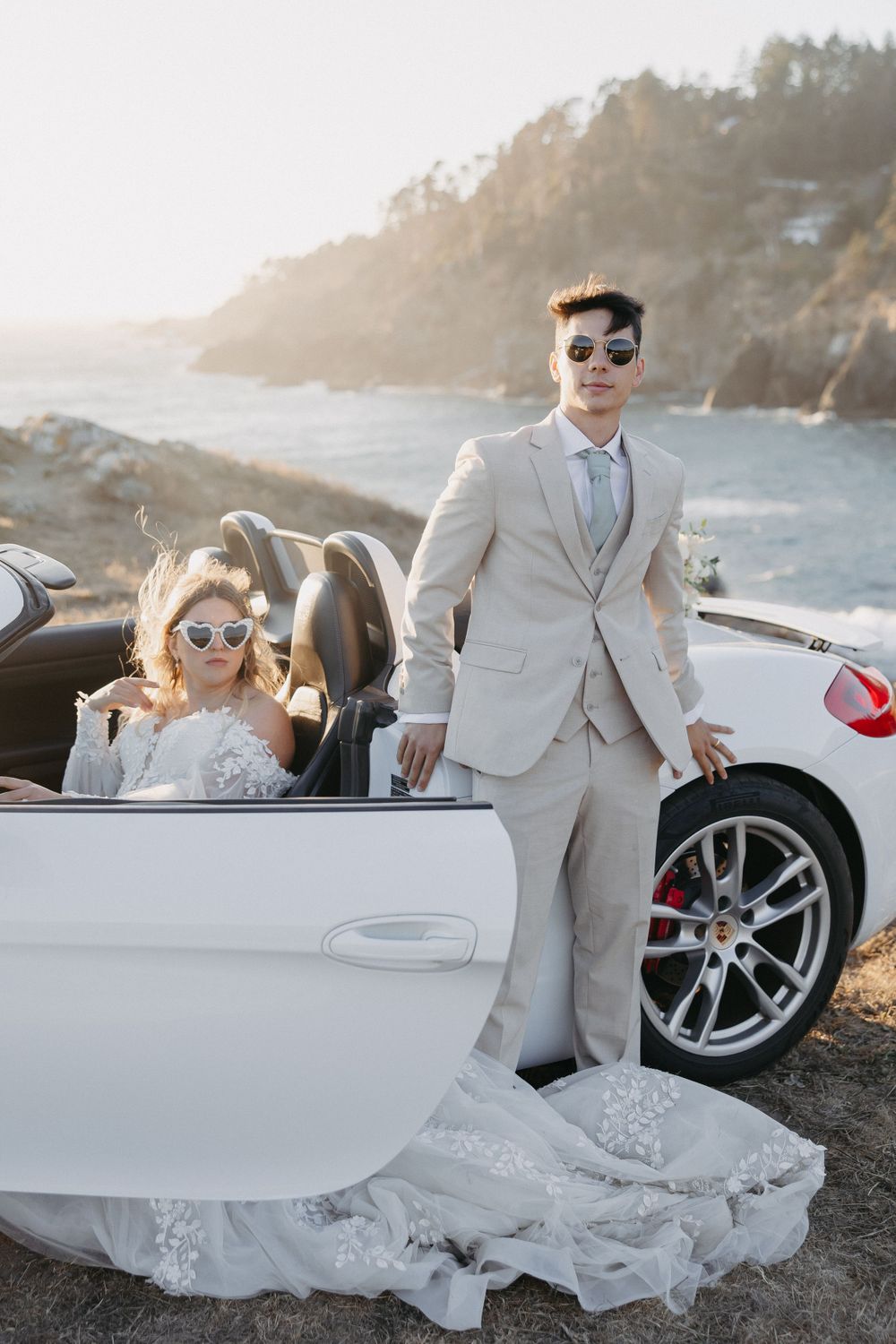 Stylish bride and groom wearing sunglasses pose with their white convertible sports car.