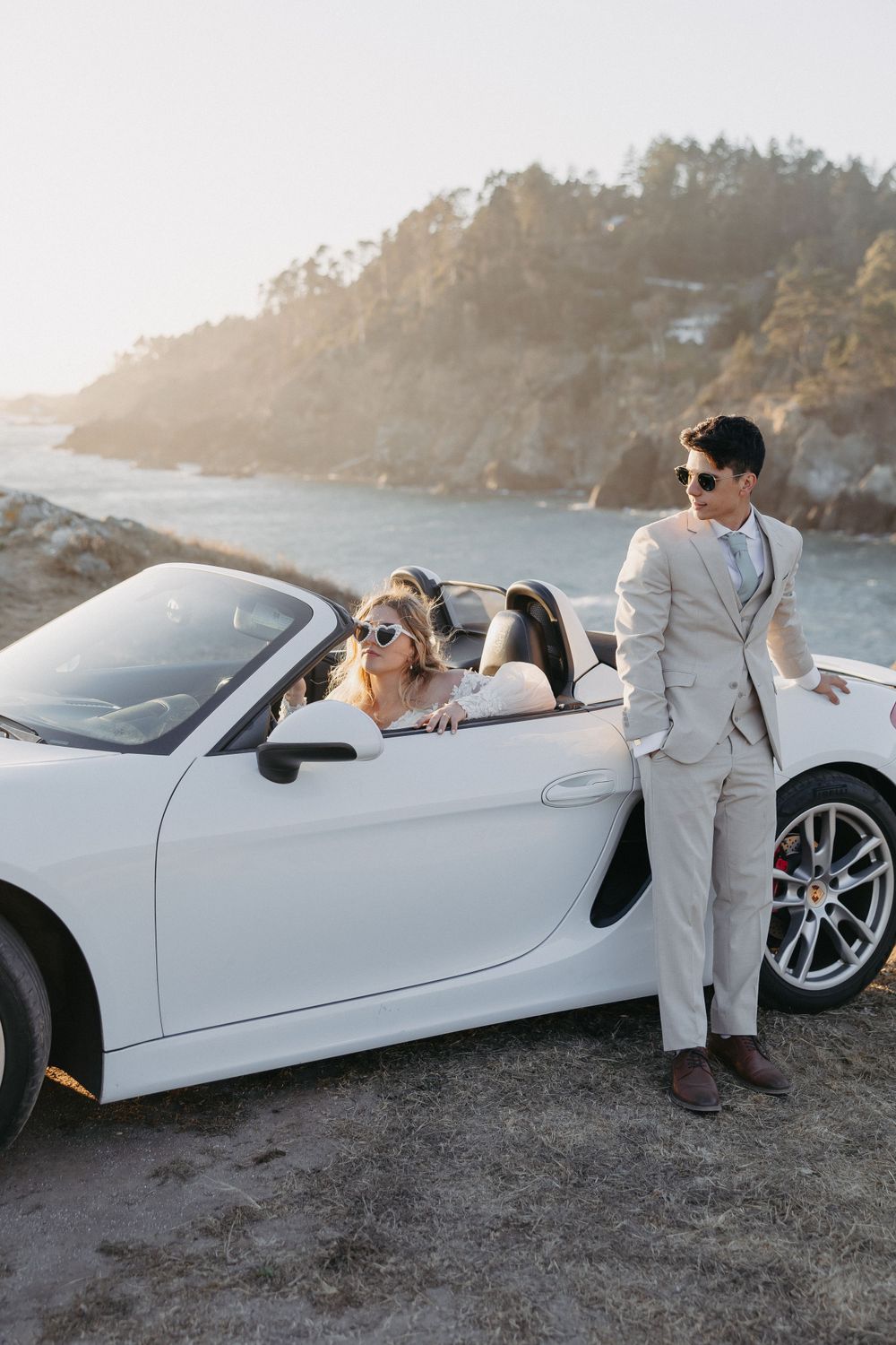 Stylish wedding couple posing with their white convertible sports car on a scenic coastal overlook.