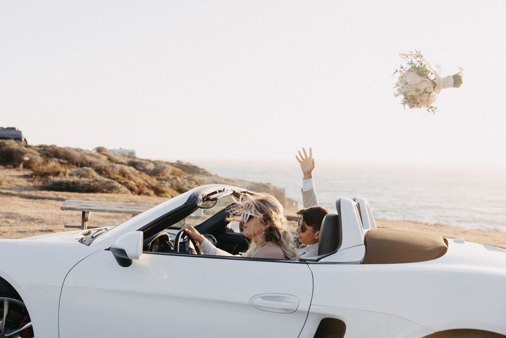 Person in white convertible car throws bouquet while driving along coastal road with ocean view.