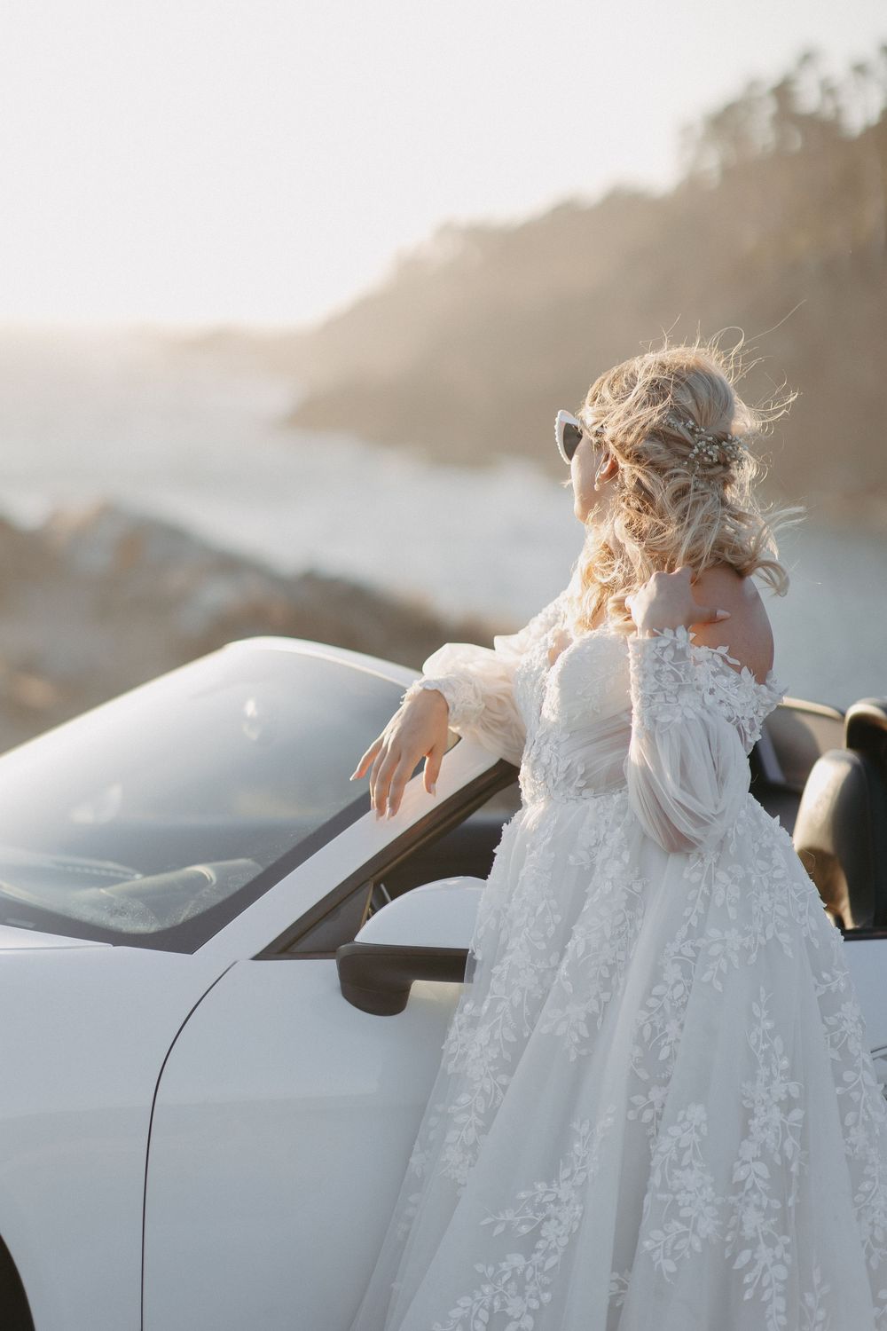 A bride in a lace gown stands by a white sports car as her hair flows in the coastal breeze.