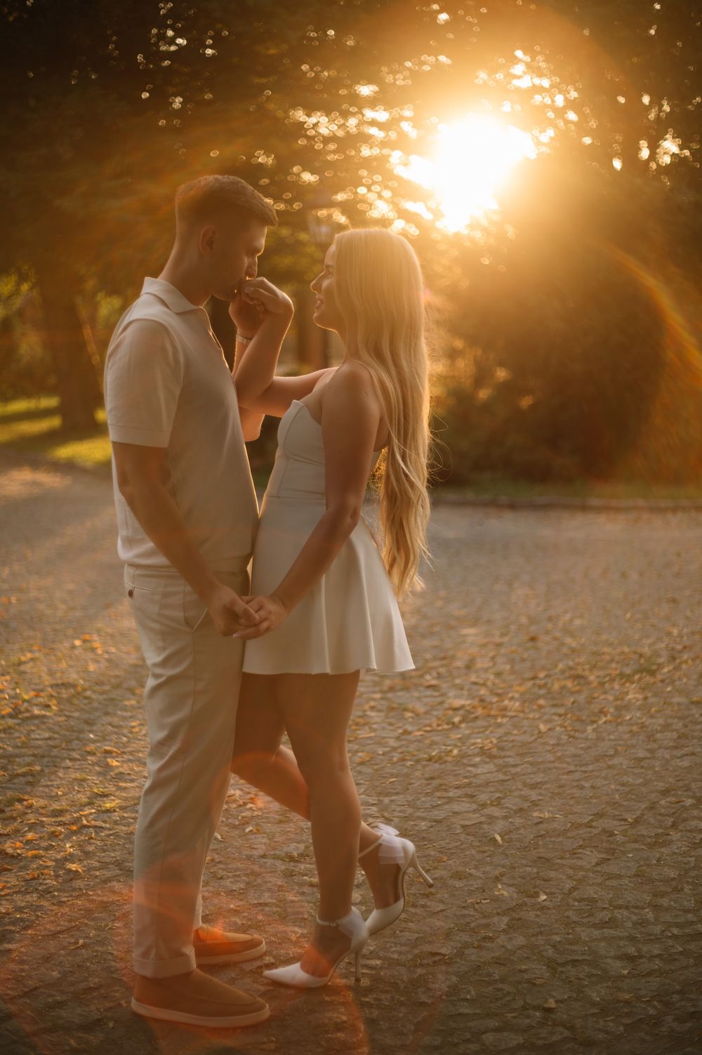 A romantic couple shares an intimate moment at sunset on a dirt path with golden sunlight streaming through trees.