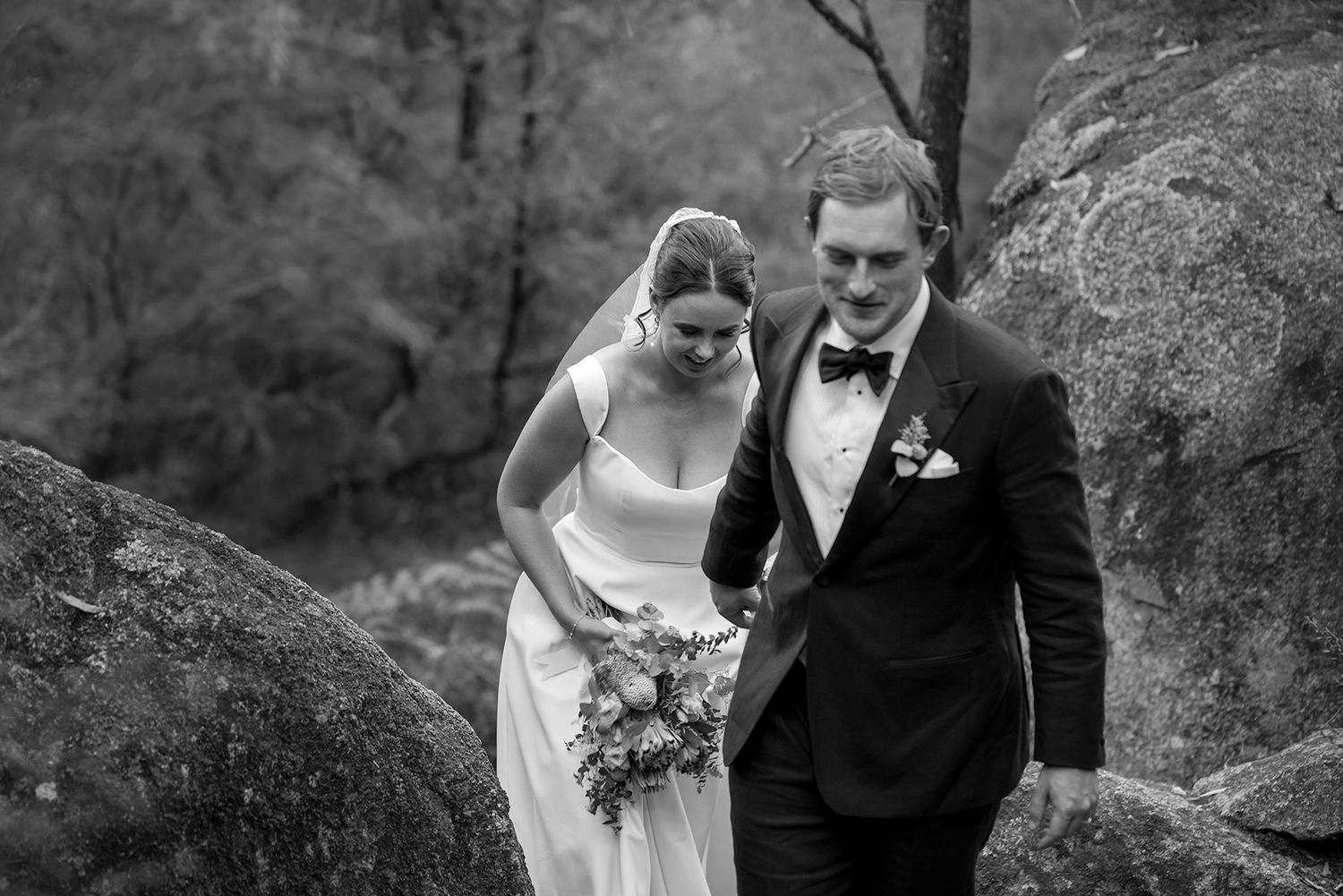 A bride and groom walk together between large rocks outdoors in a black and white photograph.