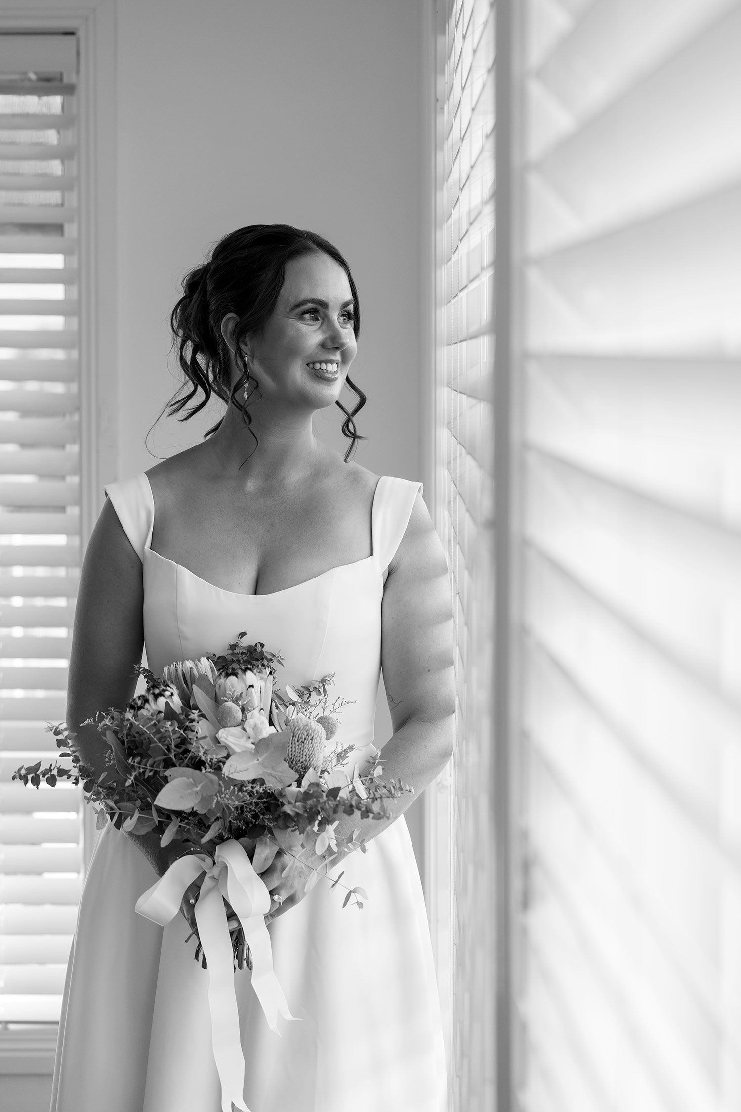 Black and white portrait of a bride in a sleeveless white dress holding a floral bouquet by a window with shutters.