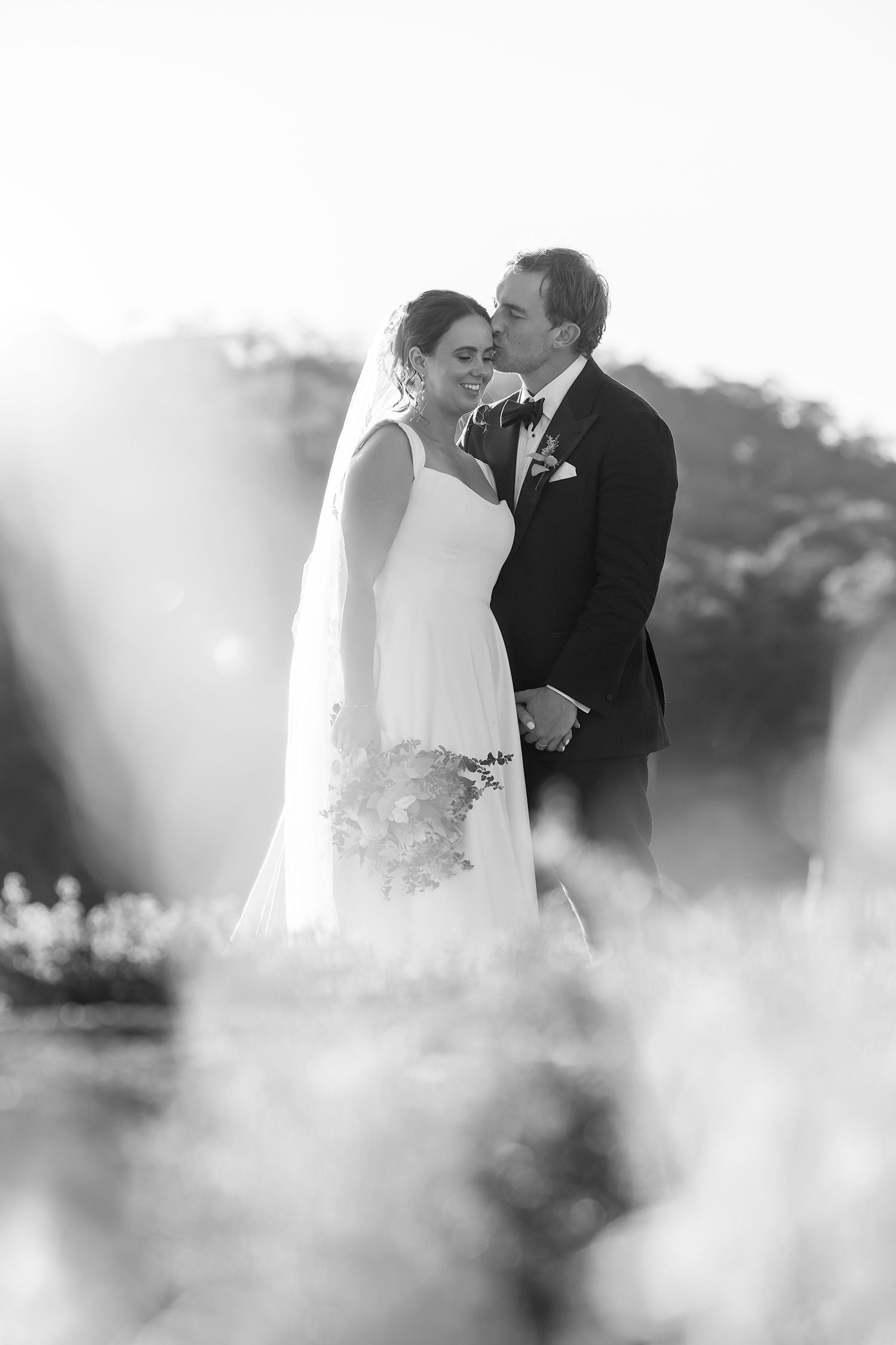 Black and white wedding photograph showing a romantic moment between newlyweds in an outdoor setting with soft focus foreground.