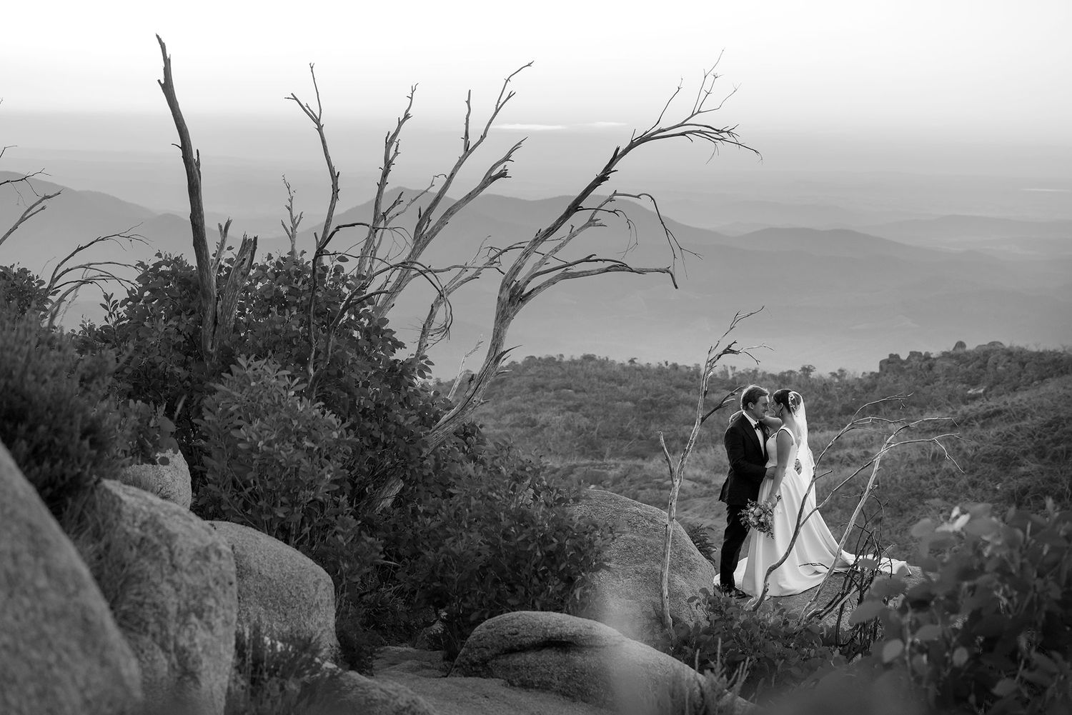 A couple stands among boulders and bare trees on a mountaintop during sunset with misty hills in the distance at the Horn Mt.Buffalo.