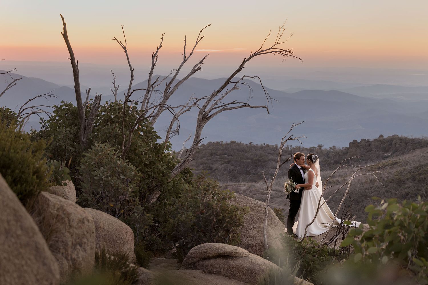 A couple stands among boulders and bare trees on a mountaintop during sunset with misty hills in the distance at the Horn Mt.Buffalo.