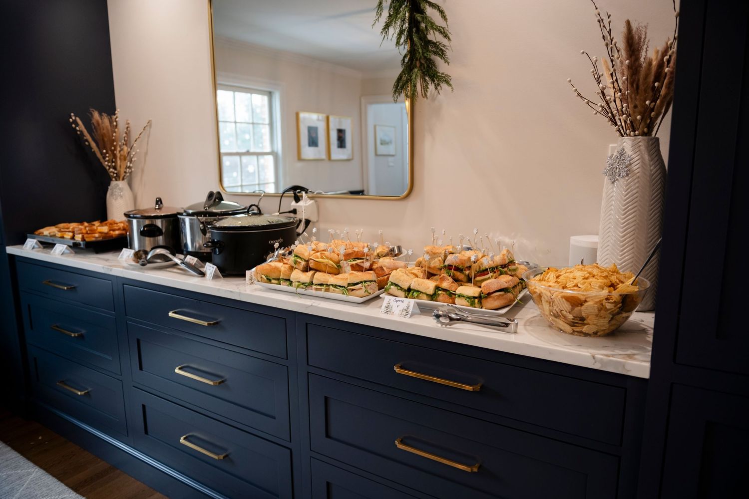 A buffet spread of sandwiches and appetizers arranged on a navy blue cabinet with gold handles and white countertop.
