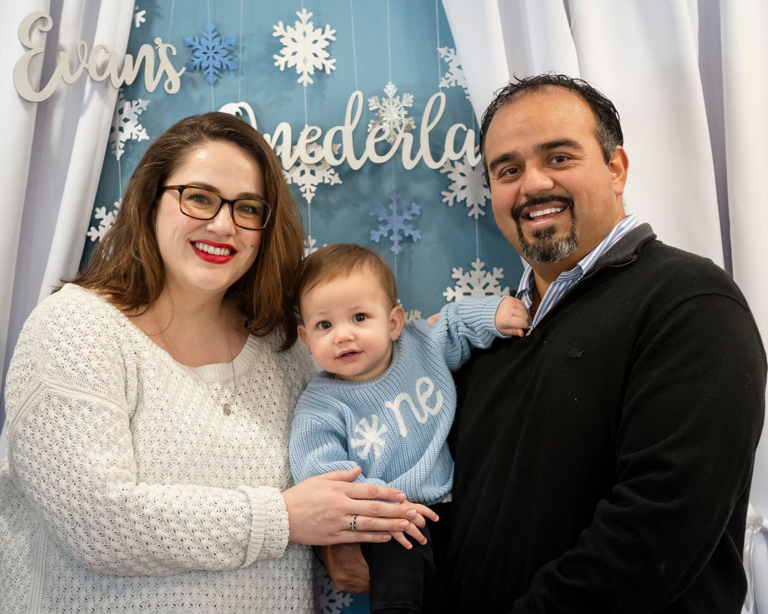 A family poses in front of a snowy winter-themed backdrop with decorative snowflakes and white curtains.
