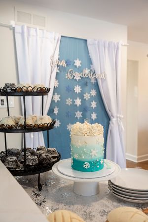 Winter theme dessert table with ombre blue cake and snowflakes against a blue backdrop with white curtains and treats display.