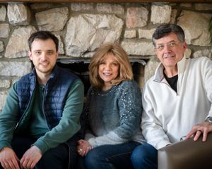 Three people sit together and smile on a couch in front of a stone fireplace.