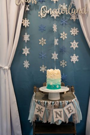 Winter themed first birthday cake display with hanging snowflakes and a blue ombre cake on white stand with ONE banner.