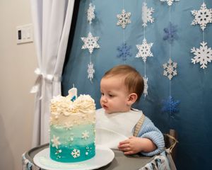 A toddler sits in front of a winter-themed cake with blue ombre frosting and snowflake decorations hanging on the wall.