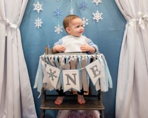 Winter-themed first birthday party setup with high chair decorated in grey and white banners and hanging snowflakes.