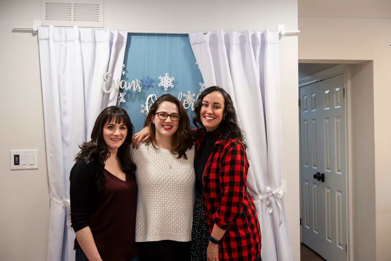 Three people stand together smiling in front of white curtains and a blue wall at an indoor gathering.