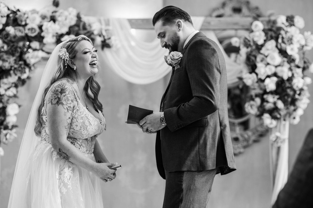 Black and white photo of a joyful wedding ceremony moment with couple laughing during their vows at floral arch.
