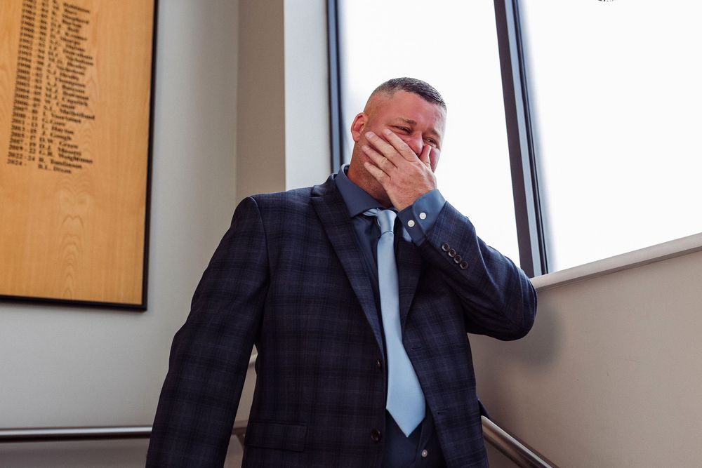 Business professional in plaid suit and blue tie covers face with hand while standing near window in office setting.