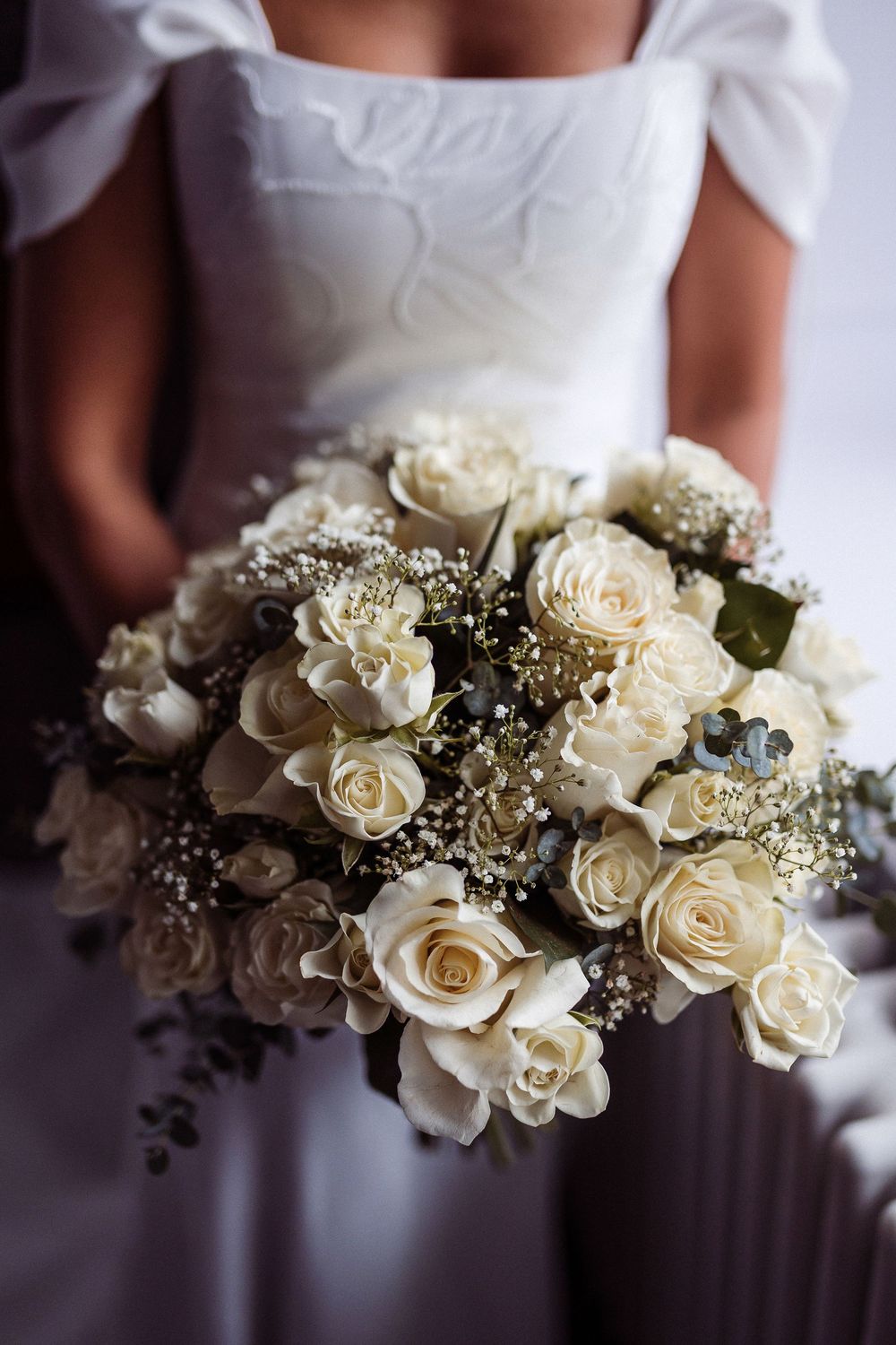 A classic white rose and baby's breath wedding bouquet held against a white dress in soft natural lighting.